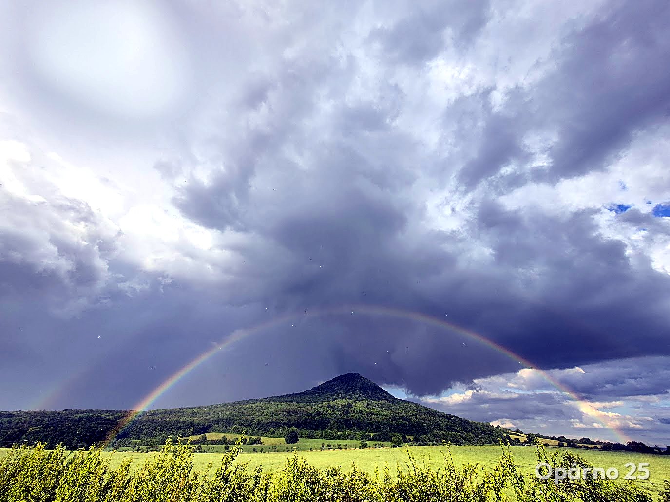 Lovoš mountain with a rainbow beneath storm clouds and green fields in the foreground.